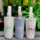 Three canvas wine totes displayed outdoors on a wooden surface, surrounded by lush tropical leaves. The totes come in three colors, natural with teal print, gray with white print, and natural with black print, each featuring the "aloha INSIDE" logo and holding wine bottles.