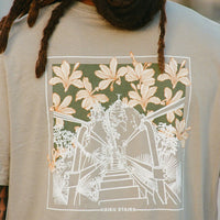 Person wearing a beige t-shirt with a detailed back design featuring an illustration of the Haiku Stairs in white, framed by tropical foliage and overlaid with large orange lilies on a dark green background. The text "HAIKU STAIRS" is printed at the bottom of the design.