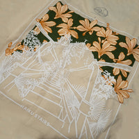 Close-up view of the back design on a beige t-shirt, featuring a white line illustration of the Haiku Stairs surrounded by tropical plants. The top portion is overlaid with large orange lilies set against a dark green background. The text "HAIKU STAIRS" appears at the bottom of the design, with a small circular logo printed above the graphic near the neckline.