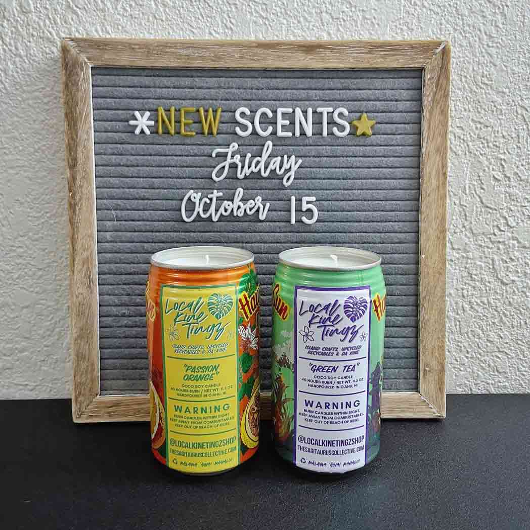 Two handcrafted soy candles in recycled Hawaiian Sun cans, Passion Orange and Green Tea, announce new scents on a letter board.
